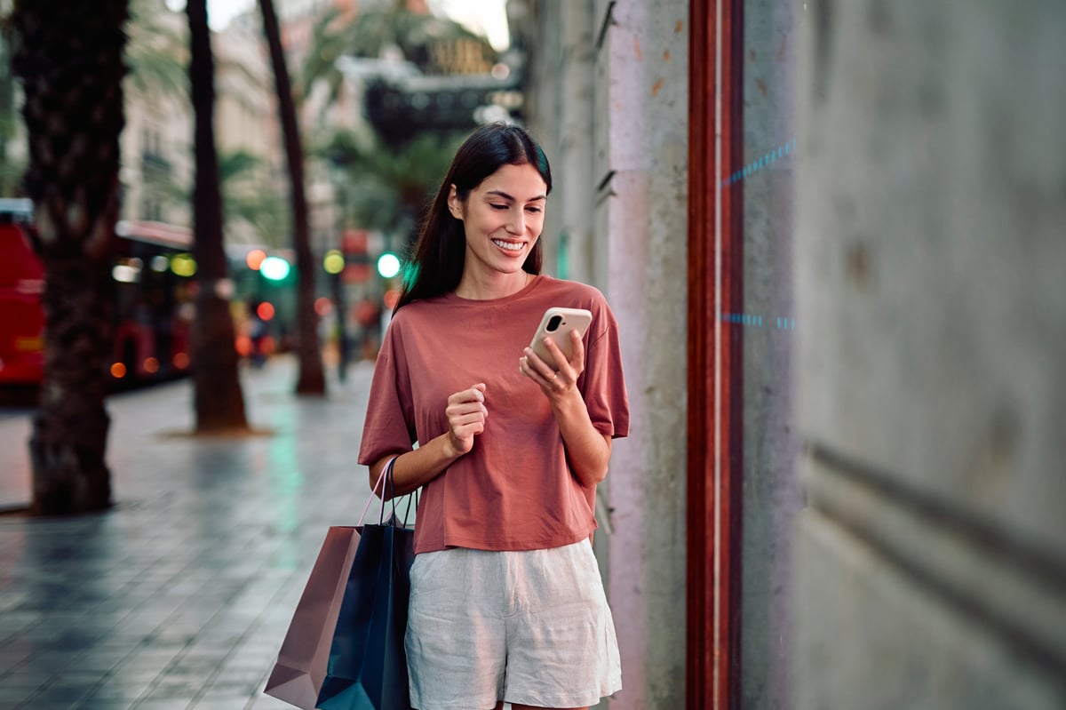 Woman smiling on a city street carrying shopping bags and checking her phone